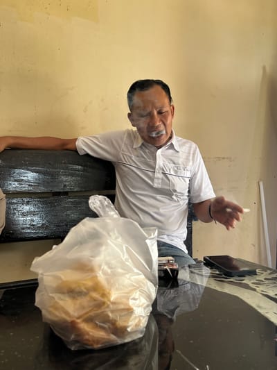 In Tanakawu, Indonesia, a man in a white polo shirt is seated on a dark wooden bench, exhaling smoke from a cigarette. A plastic bag containing food items is placed on a black, reflective table in front of him. A smartphone and a pack of cigarettes are also visible on the table. The background consists of a yellow-painted wall with visible scuff marks and a white vertical object. The lighting suggests an indoor environment with natural light.
