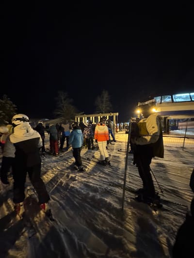 A queue of skiers waits near a lit ski lift at night. They are lined up on the snowy ground, wearing ski gear, including helmets and warm jackets. The ski lift structure is visible above them. A few bare trees stand nearby. The ground is covered with snow. It is dark in the background.