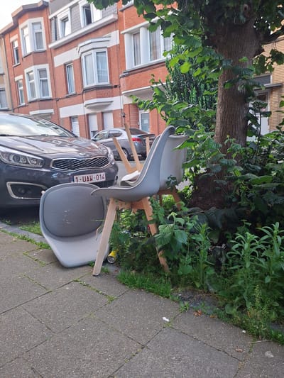 The image captures a street scene in Evere, Belgium, featuring two chairs placed precariously near a tree and a dark grey Kia car. One chair is lying on its side on the sidewalk, while the other is upright, with its back leaning against the tree. Both chairs are grey with light-colored wooden legs. The Kia car, with license plate "1-UAW-014," is parked on the street. The background shows a multi-story brick building with white-trimmed windows. Lush green foliage surrounds the chairs and the base of the tree, with some visible debris on the ground. The lighting suggests it is daytime.