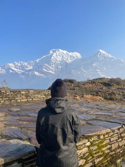 A person in a black jacket and knit cap stands on a stone path, overlooking a snow-capped mountain, with a clear blue sky above, in Lwang Ghalel, Nepal.