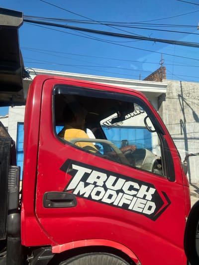 The image shows the side of a red truck with the words "TRUCK MODIFIED" in white block letters within a black arrow shape. A person wearing a yellow shirt is visible inside the truck, holding the steering wheel. The truck is parked or driving outdoors, with a clear blue sky above. Overhead power lines are visible against the sky. In the background, there are buildings with white walls and blue doors, as well as some brick structures. The overall setting appears to be an urban or semi-urban environment in Ngrame, Indonesia.