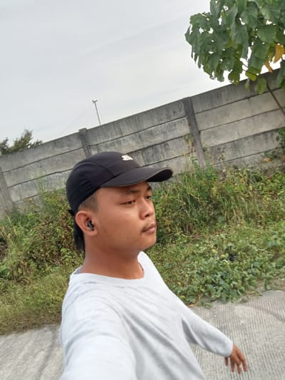 An Indonesian male wearing a gray long-sleeved shirt and a black baseball cap is captured in a side-profile, close-up shot. He is outdoors, walking on a paved path next to a concrete barrier that stretches horizontally across the frame. Lush green vegetation, including tall grasses and leafy plants, grows behind the barrier and to the right of the path. The sky above is overcast and pale. The man has short dark hair and is wearing a black earbud in his left ear. His gaze is directed forward and slightly to his right. The scene appears to be an urban or suburban environment in Teluk Kenidai, Indonesia.