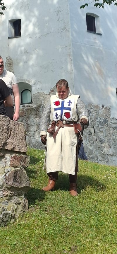 A person dressed as a medieval knight stands on a grassy area in front of a stone wall and a white building. The knight wears a chainmail shirt, a white surcoat with a blue cross and red embellishments, brown leather boots, and pouches attached to a belt. The person is looking down. In the background, there is a white building with small windows. The location is the grounds of Turku Castle in Turku, Finland.