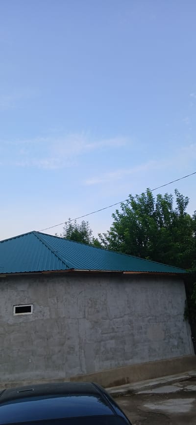The image depicts a slightly overcast sky with a few wispy clouds over a residential structure in Yangiyul, Uzbekistan. The structure features a corrugated metal roof of a teal color, a gray concrete wall with a small, rectangular, unglazed window opening, and a darker colored vehicle partially visible in the foreground. Lush green trees are situated behind the house, and a dark electrical wire stretches horizontally across the upper portion of the frame. The ground appears to be a mix of dirt and concrete. The scene suggests a quiet, daytime setting.