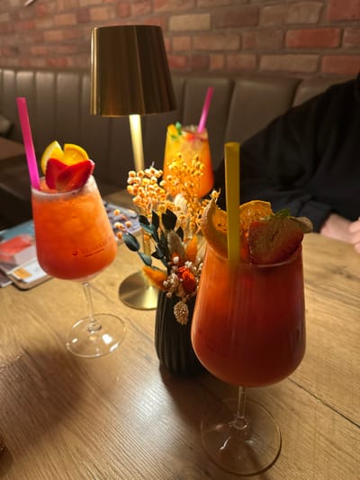 This image captures an indoor scene at a restaurant or bar in Weißenburg in Bayern, Germany. The foreground features a light-colored wooden table displaying three cocktails and a decorative centerpiece. On the right, a stemmed glass contains a red-orange beverage with a yellow straw, garnished with a dried orange slice and a strawberry half. On the left, a similar stemmed glass holds an orange-pink beverage with a pink straw, garnished with an orange wedge and a strawberry. Both stemmed glasses bear a vertical "TRINKKARTEN" inscription. Between these drinks, a small black fluted vase contains dried floral elements in orange, yellow, and muted green. A brass-toned table lamp with a cylindrical shade stands behind the floral arrangement, casting light. In the background right, a person's torso and arms are visible, clad in a dark, long-sleeved garment. Behind the table lamp, a third, smaller yellow-orange cocktail with a pink straw and a green garnish is present. The background consists of a textured brick wall and dark upholstered booth seating.