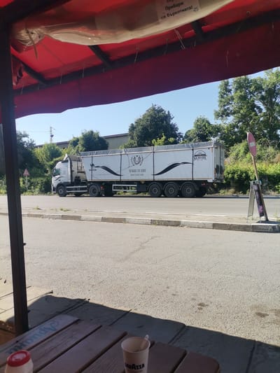 This image captures a daytime scene from beneath a red awning, likely part of an outdoor seating area. In the foreground, a wooden table is partially visible with a red-capped bottle and a paper cup. The background features a paved road with a white truck with a large trailer driving on it. The trailer is white with black graphics and text, including "MAGIC FLAME" and "KLAS OIL EST. 2001". There are trees and vegetation lining the road, and a clear blue sky above. A red and white circular sign is visible on a pole to the right of the truck, and a sandwich board sign is placed near the pole. The overall impression is a roadside view, possibly from a cafe or rest stop, in or near the city of Dralfa, Bulgaria, indicated by a sign on the awning that reads "ПРИБЕРИ СИ БЪРКОТИЯТА!" (Pick up your mess!).