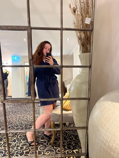 This image depicts an adult female capturing a mirror selfie within an interior space, identified as being in or near Staudernheim, Germany, based on a visible "Schloss Bad Sobernheim" tag on a decorative item. The subject, with long brown hair, is clad in a dark blue, waffle-knit bathrobe, cinched at the waist, and black slide slippers. She holds a black smartphone in her right hand, angled to capture her reflection.

The large, multi-paneled mirror, featuring an aged, grid-like frame, reflects the scene. To the left of the subject's reflection, a mannequin torso wearing a black top and patterned bottoms is visible. Further back, a green "EXIT" sign with a left-pointing arrow is mounted on a light wall, above a light fixture. Below this, a display of tall, thin bottles stands on shelves. The floor is covered by a dark carpet with a gold or beige swirling pattern.

To the right of the subject's reflection, a tall, off-white, ridged decorative vase contains dried branches and leaves, with the aforementioned "Schloss Bad Sobernheim" tag attached. A light-colored upholstered chair with wooden legs, topped by a yellow and white striped pillow, is partially visible behind the subject. Another large, ridged, off-white decorative object is partially seen in the lower right foreground. The walls are predominantly light-colored, and recessed lighting is visible on the ceiling.