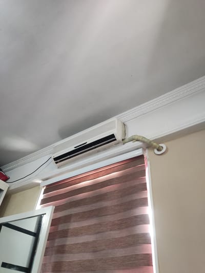 The image shows the interior of a room, likely a bedroom, in Кипчок, Tajikistan. Mounted high on the wall, just below the ceiling, is a white air conditioning unit. A white cable extends from the unit and curves to the right, disappearing behind a decorative ceiling molding. To the right of the air conditioner, a pipe wrapped in light-colored tape runs from the unit and exits through a circular white fixture on the wall. Below the air conditioner and molding, a window is covered by striped roller blinds, with light filtering through the gaps between the stripes. A portion of a white framed mirror or picture frame is visible on the left side of the image, reflecting the ceiling and air conditioner. The walls are painted a light beige color, and the ceiling is white with decorative molding. The overall impression is of a domestic interior.