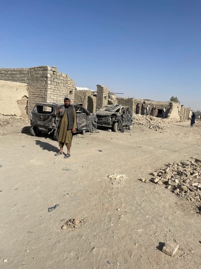 The scene depicts an outdoor, arid environment in Spin Boldak, Afghanistan, characterized by significant structural damage and debris under a clear blue sky.
In the foreground, a single adult male stands centrally, facing the camera. He wears a light brown tunic (shalwar kameez) with a dark vest, a light-colored cap, and open-toed sandals. He has a dark beard and mustache, and his right arm is slightly raised. The ground beneath him is uneven, packed dirt with scattered small rocks and tire tracks.
Immediately behind the man are two heavily damaged and charred vehicles, appearing to be SUVs or vans. The vehicle to the left is dark-colored, with severe impact damage to its rear and side, and visible burning. The vehicle to the right is also extensively damaged, with its front end crushed, hood missing, and interior elements exposed, indicating severe collision or explosive force.
In the midground and background, a row of partially collapsed single-story buildings, constructed from light-colored bricks or mud bricks, lines what appears to be a street or pathway. The structures exhibit crumbling walls, exposed interiors, and missing roof sections, with large piles of rubble and debris accumulated around their bases.
Further down the pathway, to the right, a group of approximately five to seven adult male individuals, dressed in traditional attire, are gathered near one of the damaged buildings. Their specific actions are indistinct due to distance, but they appear to be observing the area. Another lone male figure is visible walking further in the distance to the far right. The horizon in the background shows a dusty haze and a few distant trees.