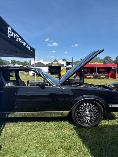 In Fairburn, United States, a black custom car is parked on a grassy field under a clear blue sky with scattered clouds. The car's hood is open, revealing the engine compartment. The car features a chrome trim along its lower body and a custom wheel with a complex silver spoke design.
Behind the car, several tents are set up. To the left, a black tent with "BitFortune.com" printed in white is visible. Further back, a large red tent with "MECUM AUCTIONS" in white lettering is prominent, with people visible behind a table. Other tents and vehicles are present in the background, suggesting an outdoor event or car show. The overall environment is an open, sunny area with neatly mowed grass.