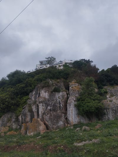 The image shows a rocky hillside topped with a building, taken on a cloudy day.

**Location:** This is likely near Tétouan, Morocco, evidenced by the building sign which reads "Café Stitika".

**Subjects:** The main subjects are the rocky terrain, vegetation, and the building at the top. The vegetation consists of green bushes and some sparse trees.

**Objects:** The building, the sign "Café Stitika", rocks, bushes and power lines.

**Environment:** The sky is overcast. The vegetation suggests a natural, possibly coastal environment. The presence of the building implies some human development.