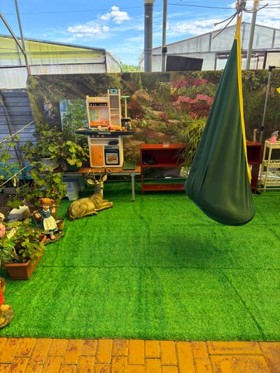 In the city of Valdgeym, Russia, an outdoor play area features a vibrant green artificial turf surface. To the left, a decorative figure of a girl carrying a basket stands near potted plants. Adjacent to her is a realistic-looking statue of a deer, lying down. Behind these figures, a wooden table supports a child's play kitchen with various accessories. A large, dark green swing hangs from a rope tied to a structure, partially obscuring a colorful tapestry depicting a garden scene. The background includes greenhouses with translucent roofs and a bright blue sky with scattered clouds. The foreground shows a yellow brick pathway bordering the artificial turf.