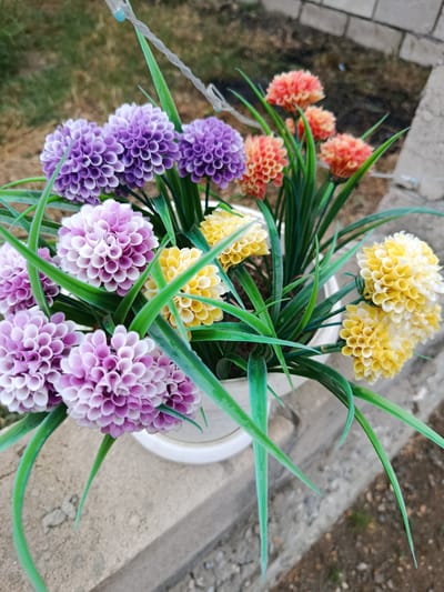 This media file features a close-up, high-angle shot of a white planter filled with artificial flowers and greenery. The flowers are arranged in clusters and come in three distinct colors: purple, yellow, and a reddish-orange. The purple flowers are prominent in the foreground, with some having lighter, almost white tips. The yellow flowers are clustered towards the center and right side of the planter. The reddish-orange flowers are visible in the background, appearing slightly out of focus. Long, bright green artificial leaves or stems emerge from the planter, creating a lush visual effect.

The planter itself is white and partially visible. It rests on a rough, textured concrete surface, possibly a ledge or wall, which is also visible in the foreground and to the right. Behind the planter, the background is blurred, showing hints of dry, brown ground and a brick wall or structure. The overall scene suggests an outdoor setting. The lighting appears to be natural, with some shadows present, hinting at it being daytime. There is no visible text in the image, and no people or animals are present. The focus is solely on the artificial floral arrangement and its immediate surroundings. The image conveys a sense of artificial vibrancy and decorative placement, likely intended to add color to an outdoor space. The location context provided is Khasavyurt, Russia.
