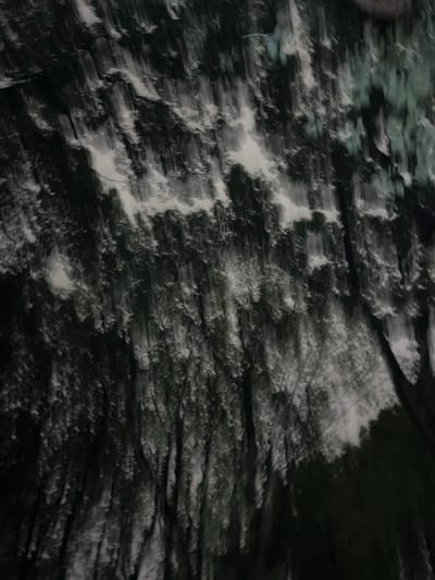 The scene, located in the city of Yanqiao, China, presents a heavily motion-blurred image dominated by vertical streaking. Dark, elongated forms, consistent with tree trunks and dense foliage, occupy the majority of the frame, appearing as vertical bands of deep green and grey. These dark elements are interspersed with irregular, lighter streaked areas of white and light grey, suggesting an overcast sky or bright light sources filtered through a canopy. In the upper right quadrant, a faint, thin, horizontally-oriented streak of turquoise light is discernible. No distinct individual objects, structures, people, or discernible actions or interactions are visible within the blurred environment.