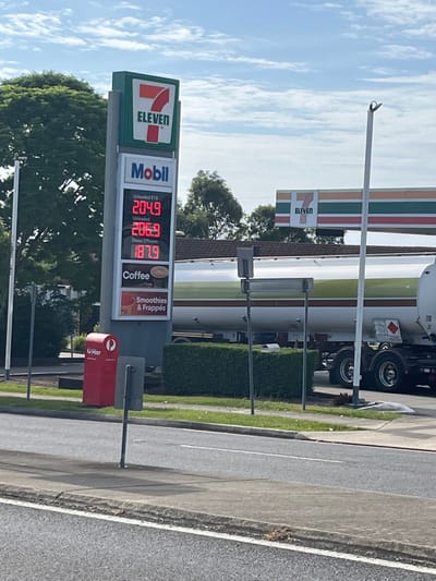 An outdoor, landscape-oriented photograph depicts a gas station and a street scene in Riverhills, Australia, under a partly cloudy sky during daylight hours. The primary focus is a tall price sign for a Mobil gas station co-located with a 7-ELEVEN store.

The sign prominently features the 7-ELEVEN logo at the top and the Mobil logo below it. Digital readouts display current fuel prices: Unleaded E10 at 204.9, Unleaded at 206.9, and Diesel Efficient at 187.9. Sections beneath the prices advertise "Coffee" and "Smoothies & Frappés".

In the background, a large fuel tanker truck, likely branded Mobil, is visible near the gas station building, which also displays a 7-ELEVEN logo. A well-maintained green hedge separates the station from the roadside. In the foreground, a red Australia Post mailbox, marked "POST," stands near the edge of a paved road. The road features a white line, a curb, and a grassy verge. Trees are scattered throughout the background, with scattered white clouds visible in the blue sky. The fuel prices and the red mailbox are indicative of an Australian location.