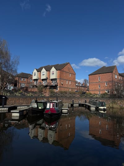 The scene depicts a canal or marina in Stourport-on-Severn, Wyre Forest, United Kingdom, under a predominantly clear, deep blue sky with scattered white cumulus clouds. Three narrowboats are moored to wooden floating pontoons on calm, dark water, which provides clear reflections of the sky and adjacent structures. The central narrowboat features a red bow section, a dark green cabin with white-framed windows, and a dark hull. To its immediate left, another narrowboat exhibits a dark hull and a green cabin. A third narrowboat, with a light green hull, is partially visible on the far right, moored to a separate pontoon.
Along the elevated bank behind the boats, a dark-colored, possibly metal, fence or railing spans a brick wall. Beyond this barrier, a series of three-story residential buildings constructed of red brick with dark brown or grey roofs are visible. The central grouping of these buildings prominently features three gabled dormers with light-colored (likely white or light grey) cladding and dark-framed windows. These buildings also include multiple balconies with dark railings. Additional brick buildings are visible to the left and right. Sparse, bare deciduous trees are present on the left bank and behind the rightmost building. No individuals are present, and no discernible activity is observed.