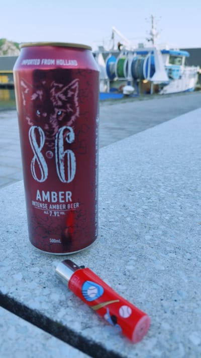 An outdoor, eye-level, medium shot captures a red 8.6 Amber beer can and a red lighter with baseball motifs resting on a textured, light-colored concrete surface. The can is positioned vertically and slightly to the left of the frame, displaying a wolf's face design and text including "IMPORTED FROM HOLLAND", "8.6 AMBER", "INTENSE AMBER BEER", "ALC 7.9% VOL", and "500mL". The lighter lies horizontally to the right of the can, its designs partially visible. In the background, slightly out of focus, a white and blue boat with fishing equipment is docked near a yellow structure. The sky above is clear and light blue. The scene appears to be at a harbor, possibly in Cherbourg-en-Cotentin, France, given the presence of a docked boat and open water. There are no people visible in the image.