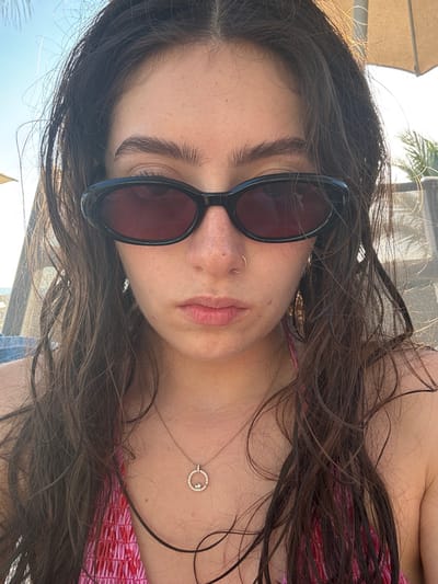A young woman with wavy brown hair and freckled skin is lying down, wearing black sunglasses. She has a nose ring and a delicate necklace with a circular pendant. The woman is wearing a pink and red top, and a beach umbrella is visible in the background. The overall impression is relaxed and summery.