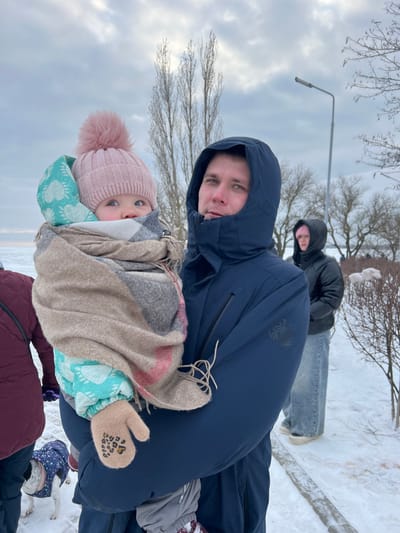 This image captures an outdoor winter scene in Primorka, Russia. In the foreground, a Caucasian man with a neutral expression, wearing a dark blue hooded winter jacket, holds an infant. The infant, wrapped in a multi-colored plaid blanket over a teal and white paw-print snowsuit, wears a pink knitted beanie with a large pom-pom and a light brown mitten with a leopard print heart design on one visible hand. The child's eyes are wide, looking slightly upwards.

To the man's right, in the mid-ground, another individual in a black hooded jacket and light blue jeans stands on a snow-covered path. On the far left, a person in a dark red jacket and a small dog wearing a patterned winter coat are partially visible.

The environment is characterized by snow-covered ground. In the background, tall, bare deciduous trees stand against an overcast sky with grey clouds. A street lamp is visible behind the man's right shoulder. Beyond the trees and snow, a body of water or ice extends to the horizon.
