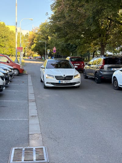In the city of Brno, Czechia, on a street lined with trees and buildings, a white Skoda Superb is parked facing the viewer. The car has its headlights on, illuminating the asphalt road. The license plate reads "RMX 09291". Several other cars are parked or driving nearby, including a red Skoda, a gray SUV, and a red van. The scene appears to be a residential or commercial area, with streetlights and traffic signs visible. The overall atmosphere is calm and ordinary, depicting a typical urban street scene.
