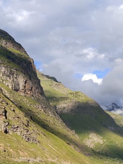 This is a vertical, medium-shot photograph of a steep, mountainous landscape. The image is dominated by the imposing presence of rugged, rocky mountains covered in vibrant green grass. These mountains are bathed in dramatic sunlight and shadow, creating a striking contrast. The sky above is a mix of bright white clouds and patches of clear blue sky, suggesting variable weather conditions. The scene appears to be set in Damphug, India, based on the provided context. No people or specific man-made objects are clearly visible, giving the impression of a vast, natural, and relatively untouched environment. The overall impression is one of raw, natural beauty and the dramatic interplay of light and shadow on geological formations.