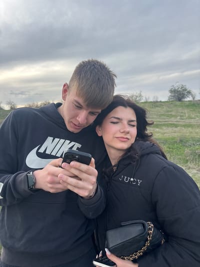 A young man and woman are lying together outdoors, with the man holding a smartphone. The man wears a dark Nike hoodie and a wristwatch. The woman is nestled against him, wearing a dark puffy jacket with the word "Juicy" on it and holding a dark, quilted purse with a gold chain. The background features a field or meadow and a cloudy sky. The location is not discernible as Tsar Asen, Bulgaria.