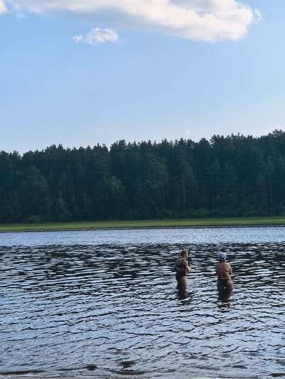 Two people stand in the shallow waters of a lake, facing away from the camera. The person on the left wears a black bikini and has blonde hair pulled back. The person on the right wears a bikini with an orange top and black bottoms and has a light-colored swim cap on. The water is rippled and reflects the sky. In the background, a dense, dark green forest lines the opposite shore of the lake. Above the forest, a light blue sky is visible with scattered white clouds. The scene appears to be set during daytime, likely late afternoon or early evening, given the soft light. The location is identified as Sysert, Russia. There is no visible text in the image.