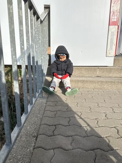 The scene depicts a young child seated outdoors in Neuhausen am Rheinfall, Switzerland, on a sunny day. The child, dressed in a black hooded winter jacket, light-colored pants, and red mittens, is positioned on a set of concrete steps. A colorful headband or cap is visible beneath the jacket's hood. Dark-colored sneakers with bright green soles are worn, and the child's feet rest on grey interlocking concrete pavers below the steps. The child's hands are clasped together in front, and their gaze is directed downwards. Behind the child is a plain white wall or structure, with a narrow vertical wooden element, partially red and white, visible to the right. To the left of the child, a metal railing, likely galvanized steel, runs vertically alongside the steps, with overgrown vegetation visible beyond it. Strong sunlight casts prominent shadows from the railing and the child diagonally across the paved ground.