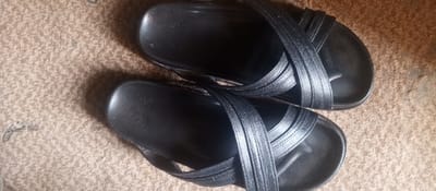 The image displays a pair of black sandals positioned on a light brown, textured floor surface, consistent with a carpet or rough-textured indoor flooring, within the city of Tunga Mekidi, Nigeria. Both sandals are oriented roughly parallel, with the left sandal slightly above and to the left of the right sandal. Each sandal features a solid black footbed and three black straps: one wide toe strap and two narrower, crisscrossing straps over the instep. The straps exhibit a slightly textured, possibly synthetic, material. Small particles or dust are visible on the surface of the footbeds and straps. The background floor shows minor discoloration and smudges, particularly in the lower left quadrant. No individuals, actions, or direct interactions are depicted.