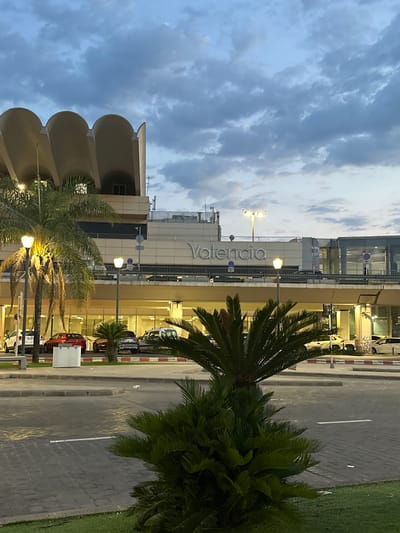 This is a full-frame, eye-level, medium shot of the Valencia Airport terminal in Manises, Spain, during the early evening. The sky is cloudy with shades of gray and blue. The airport building is visible in the background, with the word "Valencia" displayed prominently in large, silver letters on its facade. The architecture of the building features a distinctive ribbed roofline. In the foreground, palm trees and other greenery are present. Several cars are parked in a lot in front of the terminal. The scene is well-lit by streetlights and the interior lights of the airport building, casting a warm glow on the surroundings.