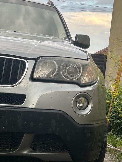 The image shows a close-up, tilted view of the front of a silver SUV, possibly a BMW X5 based on the headlights and grille design. The vehicle is parked outdoors, with a building and some vegetation visible in the background. The time of day appears to be late afternoon or early evening, indicated by the soft, diffused light and the slightly overcast sky.

The primary subject is the front of the car, focusing on the headlights, grille, and part of the bumper. The headlights are complex, with multiple lenses and circular elements. A small, circular fog light or daytime running light is also visible on the right side of the headlight assembly. The grille is a classic kidney-bean shape, typical of BMW vehicles, with chrome accents. The bumper is black and textured, with visible grilles and what appear to be parking sensors. The car is dusty, suggesting it has been driven recently or is parked in an environment where dust accumulates.

The setting appears to be a residential or semi-rural area. The building in the background has a light-colored facade and a dark-tiled roof. Green foliage, possibly from a tree or shrub, hangs down from above, adding a natural element to the scene. The sky is cloudy, with patches of lighter sky suggesting a break in the clouds.

There are no people visible in the image, and no specific activity or event is taking place. The image is purely a still shot of the vehicle. The mood is neutral, focusing on the details of the car and its immediate surroundings. There is no visible text in the image. The location context provided, Tomai, Moldova, places the scene in a specific geographical region.