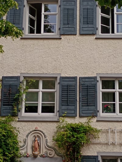 The image shows the exterior of a building with a stucco facade, likely in Meersburg, Germany. Multiple windows with dark blue shutters are visible. Some shutters are partially open, revealing the interiors. The upper windows have white frames and are divided into multiple panes. The left-most upper window is open. Green foliage from trees and vines partially obscures some of the windows and shutters. Below the windows, a decorative niche contains a statue of a woman in flowing robes. The niche is framed by an arched decorative element and flanked by floral motifs. A metal trellis structure is visible on the right side of the facade. The overall impression is of an older building with architectural detailing and a natural, overgrown aesthetic.