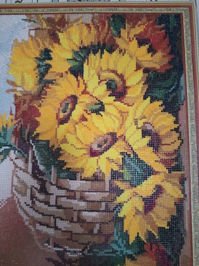 This is a close-up, eye-level shot of a framed diamond painting depicting a bouquet of sunflowers in a woven basket. The painting is rendered in a mosaic style with small, colorful diamond-shaped beads. The sunflowers are predominantly yellow with dark centers, surrounded by green leaves and accents of red and brown. The basket appears to be made of brown and tan beads, with a textured weave pattern. The background is a soft, light blue, suggesting a sky or simple backdrop. The frame surrounding the artwork is ornate and appears to be gold with intricate patterns. The image does not contain any information about the city of Älimbet, Kazakhstan.