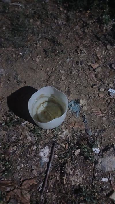 The scene appears to be a patch of dirt with debris. Visible is a white plastic container, partially filled with a murky liquid. The surrounding ground is covered with small stones, vegetation, litter, and a long, dark, thin object that may be a stick or piece of metal. The image is dimly lit, suggesting it was taken at night. There are no people or interactions visible. It is presumed to be taken in Putucual, Venezuela.