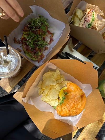 The image shows a table with three food containers and two drinks. The containers hold a sandwich with chips, avocado toast, and a croissant with chips. The drinks are in clear plastic cups, one with a black straw. A hand with a ring is visible near the avocado toast. The setting appears to be outdoors on a wooden table. The image was taken in Chula Vista, California.