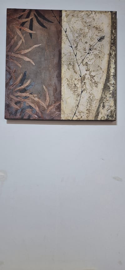 A framed artwork with a textured abstract design is hanging on a plain, light-colored wall. The artwork is divided vertically. The left half features a dark brown background with abstract, leaf-like shapes in shades of brown and grey, appearing to be applied with a textured technique. There are also several faint circular motifs on this side. The right half of the artwork has a textured, off-white and beige background with darker, more muted tones in places, creating a mottled effect. A thin, dark branch-like element with small dark dots, possibly representing buds or berries, extends diagonally from the top left to the bottom right across the right half of the artwork. There is a distinct black border framing the entire artwork. The wall below the artwork shows some minor imperfections, including light discoloration and small patches of peeling paint. The image appears to be taken in an indoor setting, likely a home or gallery. The specific location in Póvoa de Varzim, Portugal, cannot be determined from the visual information provided.