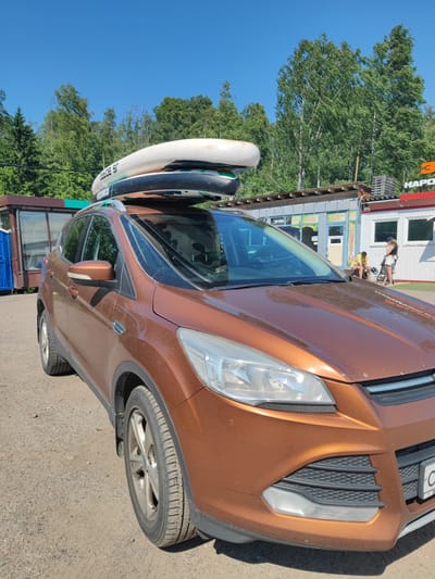 A brown Ford Kuga SUV is parked on a dirt lot outside a row of buildings. The SUV has two paddleboards strapped to its roof rack. In the background, a woman in a yellow shirt and a woman in a black top and white shorts are standing near a building with a red "HAPO" sign. The area appears to be a commercial district.