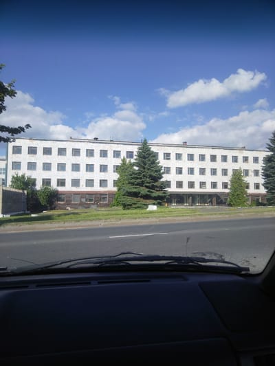 This media file shows a three-story building with a white tiled facade and many windows. The building is rectangular and appears to be an office or institutional building. In front of the building is a strip of green grass and several evergreen trees, with a road in the foreground. The sky is blue with scattered white clouds, indicating a daytime scene. The overall impression is one of a typical day in an urban or suburban environment. The image is taken from the perspective of someone inside a vehicle, looking out through the windshield. There are no people or visible text in the image. The location is specified as Vawkavysk, Belarus.
