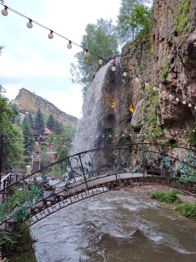 The image depicts a picturesque scene at a waterfall in the city of Krasnyy Kurgan, Russia. A decorative metal bridge, adorned with artificial greenery, spans a fast-flowing river in the foreground. The bridge's intricate railing features swirling patterns and circular motifs. Above and behind the bridge, a majestic waterfall cascades down a sheer rock face. The rock face is covered in patches of green vegetation and shows signs of weathering. A string of globe lights is suspended along the cliffside, adding a festive touch to the natural landscape. In the background, a mountainous terrain rises, with several buildings nestled on its slopes. Tall, slender evergreen trees are interspersed among the buildings. The sky is overcast with a light-colored cloud cover. The water in the river is a murky brown, with ripples and whitecaps indicating its turbulent flow. There are no visible people in the immediate vicinity of the bridge or the waterfall. The overall impression is one of a natural wonder enhanced by human artistry and decorative lighting.