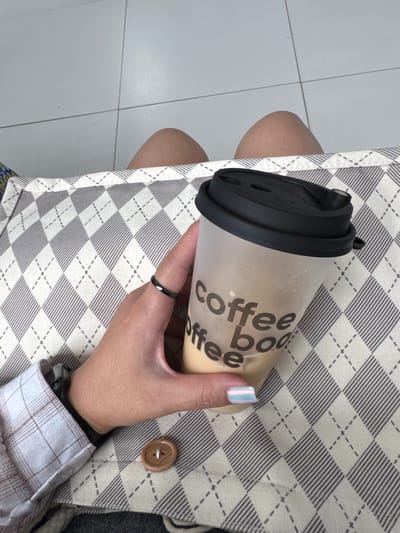 A person's hand holds a clear plastic tumbler filled with a light-colored beverage, possibly coffee with milk. The tumbler has a black lid and text printed on its side that reads "coffee boo ffee". The hand is adorned with a dark ring on the index finger and has nail polish with an iridescent finish. The person is wearing a plaid shirt, partially visible, and a watch on the wrist. The tumbler and hand rest on a surface covered with a fabric featuring a diamond pattern in shades of beige and grey. The background shows a light-colored tiled floor. The scene appears to be indoors, possibly in a cafe or a home. The image does not provide any specific information that would allow for its localization in Kyzylorda, Kazakhstan.