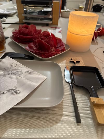 An indoor tabletop scene in Bellevue, Switzerland, is meticulously set for a festive meal, likely a raclette dinner, taking place in the evening. The soft, warm glow emanating from a cylindrical white lantern casts a cozy ambiance over the setting.
A large white serving bowl holds an abundant portion of thinly sliced red cured meat, possibly prosciutto or bresaola, elegantly arranged in ruffled layers. Adjacent to this, a light gray square plate with rounded corners awaits a diner, topped with a folded white and gray paper napkin featuring a decorative pattern reminiscent of festive baubles.
On a light-colored, woven placemat, a black individual raclette pan is positioned, accompanied by a small metal knife with a black handle. To the left, parts of a wooden raclette grill structure are visible, confirming the nature of the meal. A small black star-shaped decoration adds a subtle festive touch to the placemat.
In the background, red spherical ornaments are partially visible near the lantern, and a white card behind the meat bowl displays a partial word, "CHRISTMAS," further emphasizing a holiday or celebratory occasion. The arrangement of food, dining ware, and festive decorations strongly suggests a social gathering centered around the popular Swiss raclette, typically enjoyed during colder months or holidays. No people are visible, but the prepared scene exudes a warm and inviting atmosphere.
