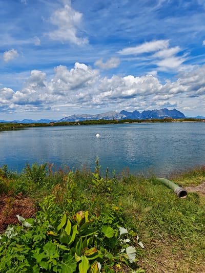 This is a landscape photograph taken on a sunny day with scattered clouds in the sky. The scene depicts a serene lake with mountains in the background. In the foreground, lush green vegetation and wild grasses line the edge of the lake. A large, green pipe emerges from the grass on the right side of the frame, partially submerged in the water.
In the mid-ground, the calm surface of the lake reflects the blue sky and white clouds. A white buoy is visible in the water. Across the lake, the distant mountains rise majestically, their peaks possibly snow-capped. Along the lakeshore in the background, several small figures of people can be seen, some walking or standing, suggesting a leisurely day out. A small building or structure is also visible on the far shore near the mountains.
The overall impression is one of natural beauty and tranquility. The bright sunlight and clear sky indicate pleasant weather, likely mid-morning or afternoon. The location is suggested to be Kirchberg in Tirol, Austria, known for its mountainous terrain and natural lakes. No visible text is present in the image.