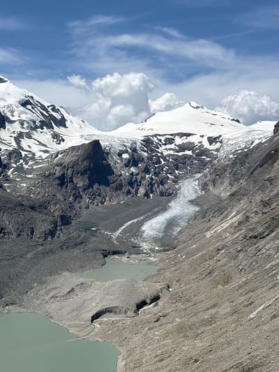 The image depicts a vast glacial landscape. In the foreground, a body of milky, turquoise water is visible, likely a glacial lake. The lake is partially surrounded by debris fields composed of rock and gravel, evidence of glacial erosion and moraines. Further up the slope, a large glacier descends, its surface characterized by crevasses and seracs. The glacier is bordered by steep, rocky mountainsides. The upper reaches of the mountains are capped with snow and ice, with dramatic peaks piercing a blue sky dotted with white, cumulus clouds. This scene is characteristic of the high alpine environment found near Heiligenblut am Großglockner in Austria, a region dominated by glaciers and rugged mountain terrain. There are no people or animals visible in the image.