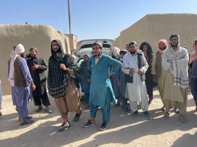 The image presents an outdoor scene in Spin Boldak, Afghanistan, under a clear, bright blue sky with strong sunlight casting distinct shadows on the unpaved, dusty ground. The background consists of several earthen-toned, adobe-style walls, likely belonging to buildings, along with a utility pole and overhead wires.
A group of approximately 12-15 adult males is visible, predominantly standing in the mid-ground. Most are dressed in traditional Afghan attire, including shalwar kameez, with some wearing vests, turbans, or scarves draped over their heads or shoulders.
Key individuals and objects from left to right include:
* A man wearing a white turban, blue shalwar kameez, and a brown vest, with his back partially to the camera, facing another man.
* A bearded man in dark clothing holding a mobile phone to his right ear.
* A man in the foreground wearing a dark plaid shawl over his head and shoulders, brown shalwar kameez, and athletic shoes, looking forward.
* A man in a teal shalwar kameez and athletic shoes, standing with his hands on his hips, facing slightly left.
* Behind these individuals, a white sedan with its trunk open is parked.
* Further right, a bearded man in a light-colored shalwar kameez and dark vest, with a white cloth draped over his left shoulder, holds a thin, dark object in his right hand.
* On the far right, a man wearing a white and dark plaid scarf draped over his head, a light brown shalwar kameez, and sandals, stands with his arms crossed, looking directly at the camera.
* Several other men are partially visible behind and between the foreground figures, observing the surroundings.