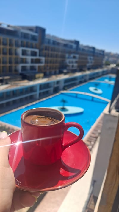 A close-up shot shows a hand holding a red coffee cup and saucer filled with dark coffee, with steam rising from the surface. In the background, a bright blue swimming pool stretches out, with white bridges and circular platforms. Beyond the pool, a multi-story building complex with a modern architectural design is visible under a clear blue sky. The scene appears to be a residential complex in Çiğli, Turkey, on a sunny day.
