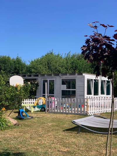 A sunny outdoor scene in Rewal, Poland, features a white wooden playhouse with large windows and a white picket fence surrounding a portion of the yard. Inside the fenced area, a yellow ride-on toy car and a small green table are visible, along with what appears to be a child wearing a pink outfit. Outside the fence, a blue toy is placed on a tire, and a large rock sits near some greenery. In the foreground, a lounge chair is positioned on the grass, and a young tree with dark leaves stands to the right. A basketball hoop is mounted on the left side of the playhouse, partially obscured by lush green bushes. The background is dominated by a clear blue sky and dense green trees.