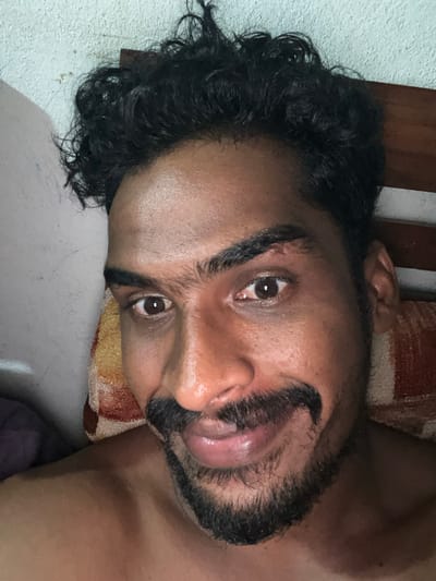 A close-up, horizontal portrait captures a man of South Asian descent lying down, looking directly at the camera. He has dark, curly hair, a full beard, and a mustache. His skin is a warm brown tone. He appears to be relaxed, with his tongue slightly extended between his lips, giving a playful or informal expression. His dark eyes are wide open, engaging the viewer.

The man is positioned on his side, with his head resting on what appears to be a pillow or cushion with a patterned fabric, featuring shades of red, orange, and yellow. Behind him, a dark wooden surface, possibly part of a bed frame or furniture, is visible. A light-colored wall or surface can be seen in the background, contrasting with his dark hair. The lighting is soft and even, suggesting an indoor setting. No specific location cues are visible within the image to indicate Ulla, Sri Lanka, other than the provided context. There is no visible text.