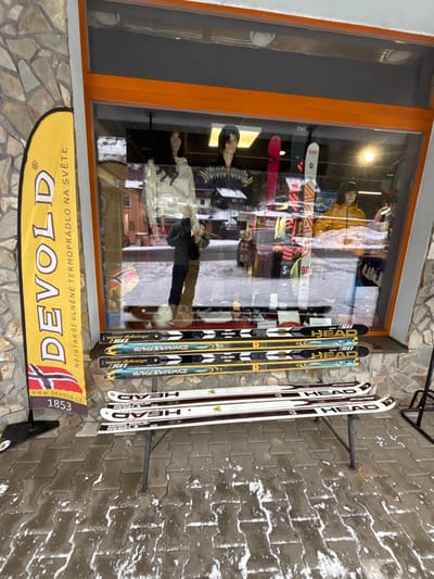 The image displays a ski shop storefront in Velká Úpa, Czechia, under winter conditions. The building features a rough-cut stone facade and a large display window framed in orange.

Inside the window, three mannequins are visible: a female mannequin on the left wearing a light-colored long-sleeve top, a central mannequin with dark hair in a patterned top, and a mannequin on the right in a yellow jacket. Several skis are displayed upright in the background, including at least two "ZAG" branded pairs. Reflections in the window show a snow-covered street, parked vehicles (one dark red/brown), and distant buildings, suggesting an outdoor urban environment. A partial reflection of a person wearing a dark coat and possibly carrying a bag is visible near the base of the central mannequin.

To the left of the window, a tall, yellow vertical banner stands on a black base. It displays the brand "DEVOLD" in large black letters, a small Norwegian flag, the text "NEJSTARŠÍ VLNĚNÉ TERMOPRÁDLO NA SVĚTĚ" (The oldest woolen base layer in the world), "WWW.DEVOLD.CZ", and "1853".

In front of the display window, six pairs of skis are arranged horizontally on a black metal folding stand. The upper layer consists of three pairs, predominantly "HEAD" and "DYNASTAR" branded, featuring designs in black, yellow, and blue. The lower layer consists of three "HEAD" branded pairs, primarily white with black and red accents, including models identified as "i.RACE REBEL".

The ground in the foreground is paved with dark gray rectangular pavers, partially covered with irregular patches of melting snow and ice.