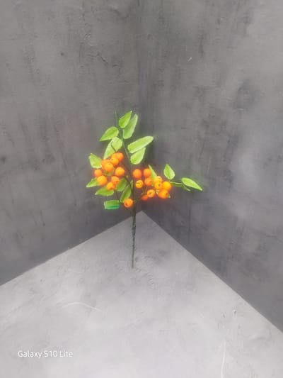 The image shows a close-up, high-angle shot of a faux berry branch with orange berries and green leaves against a dark gray, textured background. The branch is positioned in the corner formed by two walls meeting at a right angle, with one wall appearing darker and the other lighter. The lighting creates a shadow effect, with a diagonal line of light and shadow across the lower part of the image. The bottom right corner of the image features a watermark that reads "Galaxy S10 Lite". There are no people or any discernible city landmarks visible. The scene appears to be an indoor setting, possibly a studio or a display area, and there is no indication of it being located in Wellingborough, United Kingdom.
