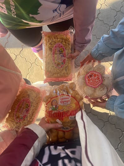 Four individuals are holding various packages of what appear to be traditional Tatar or Bashkir sweets. The sweets include "chak-chak" and "baursak". The location may be near Nizhnie Yaki, Russia, as indicated on the packaging. The individuals are wearing casual clothing. The image is taken from a top-down perspective, focusing on the sweets being held. The environment is outdoors on a patterned surface.